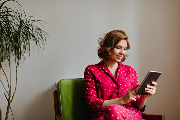 Mujer alegre sonriente en ropa de dormir con tableta digital. Filmación en interiores de mujer en pijama rojo con gadget.