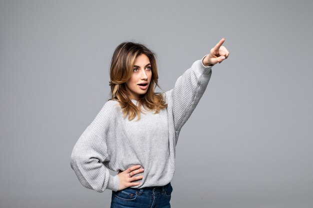 Foto gratuita mujer alegre sonriente en suéter flojo que presenta contra la pared gris del estudio que señala en el espacio de la copia para el anuncio o el texto promocional. emociones positivas, sentimientos, alegría, felicidad.