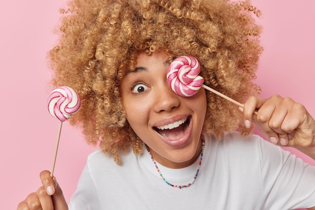 Una mujer alegre y sorprendida con el pelo rizado sostiene dos caramelos en palitos que disfruta comiendo dulces sonrisas y viste una camiseta blanca informal con fondo rosa Prueba estas piruletas