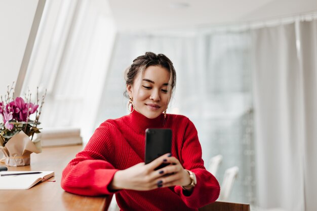 Mujer alegre en suéter de punto brillante hace selfie sentado a la mesa en una habitación luminosa junto a la ventana