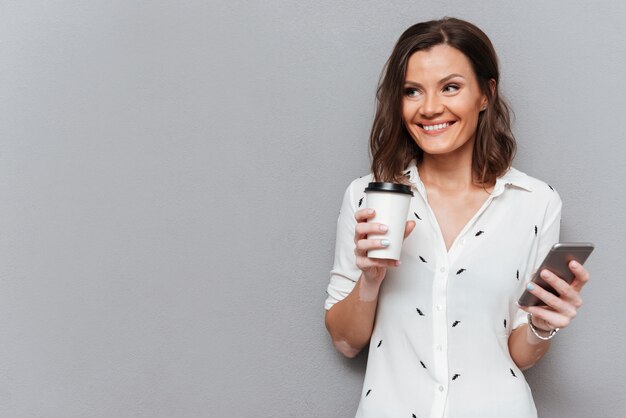 Mujer alegre con taza de café y teléfono inteligente en manos mirando a otro lado en gris