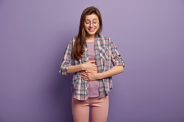 La mujer alegre tiene el estómago lleno después de una deliciosa cena, mantiene ambas manos en el vientre, está bien alimentada, sonríe positivamente vestida con ropa casual, gafas redondas aisladas sobre una pared púrpura. Sensación de saciedad