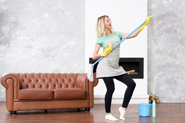 Mujer alegre tocando la fregona como guitarra de pie en casa cerca del sofá