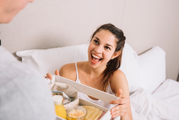 Mujer alegre tomando bandeja con desayuno en la cama