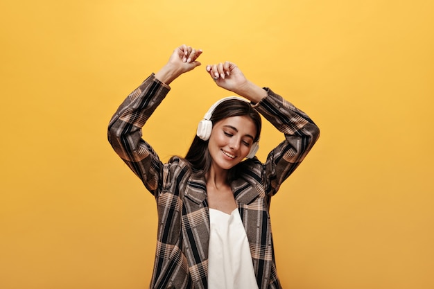 Mujer alegre en top blanco, elegante chaqueta marrón sonríe y baila en la pared amarilla