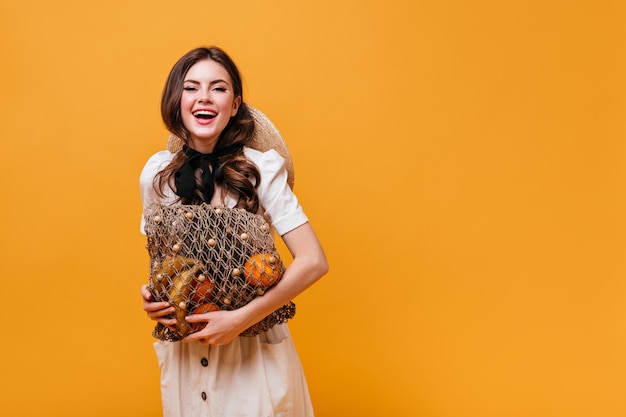 Mujer alegre en traje blanco con lazo negro sostiene bolsa de hilo con frutas sobre fondo naranja.