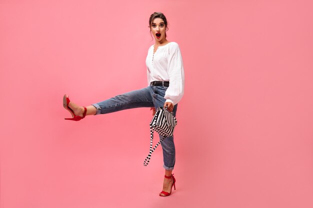 Mujer alegre en traje elegante se mueve sobre fondo rosa. Chica de moda joven con camisa blanca de labios rojos y jeans ligeros posando sobre fondo aislado.