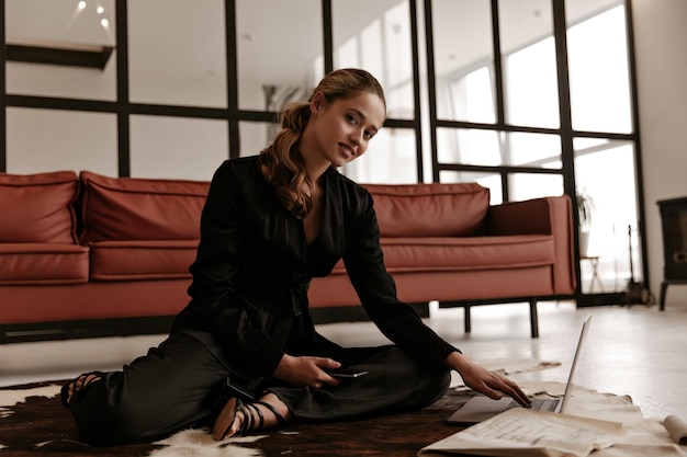 Mujer alegre en traje de seda negra de moda se sienta en la alfombra en el suelo sonríe mira a la cámara y trabaja