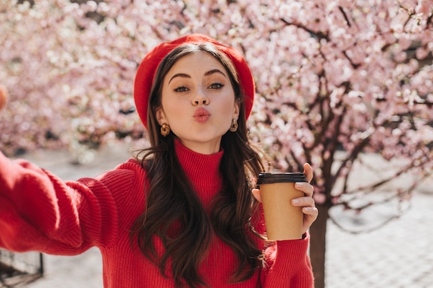 Mujer alegre con vaso de té en sus manos sopla beso y toma selfie. Retrato de dama de suéter rojo sosteniendo la taza de café contra la flor de sakura