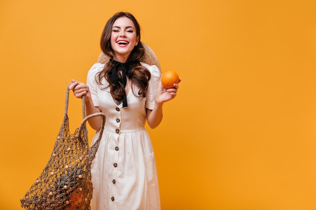 Mujer alegre en vestido blanco posando con bolsa de compras y naranja sobre fondo aislado.
