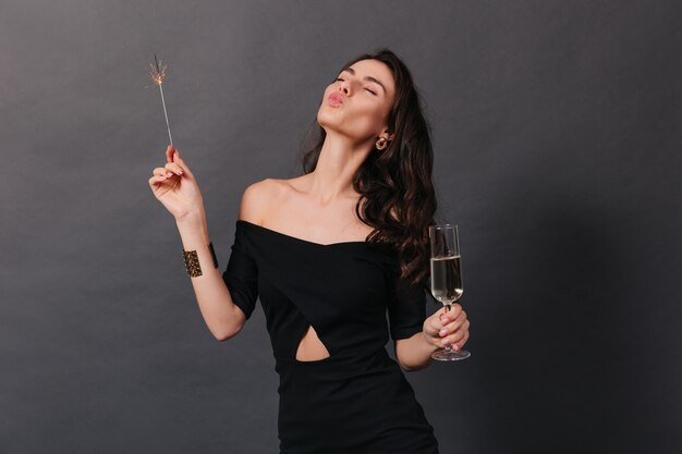 Mujer alegre en vestido de moda posando con copa de champán y bengala. La señora envía un beso sobre fondo negro.