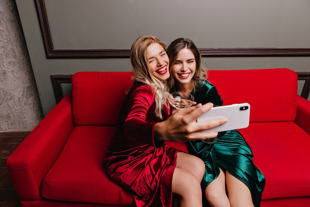 Mujer alegre en vestido rojo sentado en el sofá y haciendo sefie. Riendo a chicas en ropa elegante posando en el sofá.