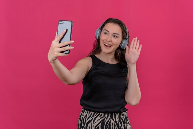 Mujer alegre vistiendo camiseta negra en auriculares habla a través del teléfono en la pared rosa