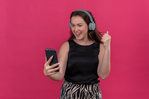 Mujer alegre vistiendo camiseta negra escucha música de auriculares y baila en la pared rosa