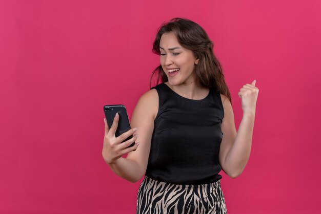 Mujer alegre vistiendo camiseta negra escuchar música desde el teléfono en la pared rosa