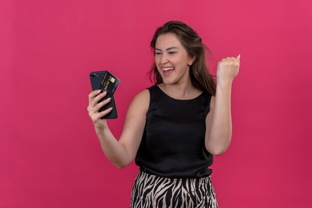 Mujer alegre vistiendo camiseta negra sosteniendo un teléfono y una tarjeta bancaria en la pared rosa