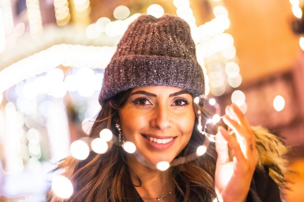 Mujer alegre vistiendo un gorro y posando mientras sostiene las luces de Navidad al aire libre
