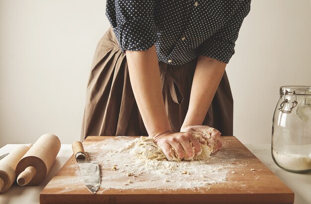Mujer amasa la masa para la pasta en una tabla de madera cerca de dos rodillos y un tarro con harina