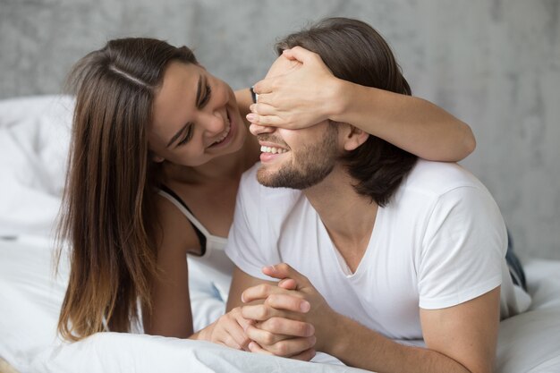 Mujer amorosa cerrando los ojos del hombre con las manos jugando en la cama