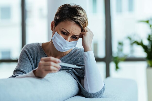 Mujer angustiada con mascarilla protectora mientras usa termómetro y mide su temperatura en casa