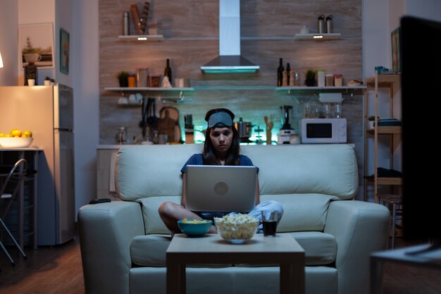 Mujer con antifaz usando laptop por la noche mientras ve la televisión y come bocadillos. Persona feliz en pijama sentado en el sofá leyendo escrito buscando navegar en el portátil usando internet comprobando correos