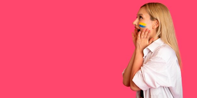 Mujer de apariencia modelo con la bandera de Ucrania en su rostro sostiene posando sobre un fondo rosa