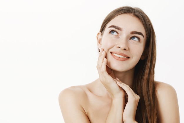 Mujer aplicando crema en la cara sintiendo la piel suave y tierna de pie soñadora y encantada con el resultado mirando la esquina superior izquierda con una sonrisa sensual tocando la mejilla posando desnuda sobre una pared gris