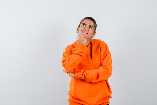 Mujer apoyando la barbilla en el puño en sudadera con capucha naranja y mirando esperanzada