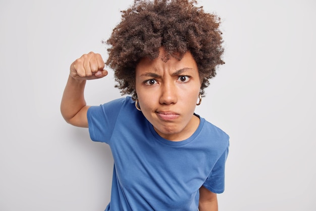 La mujer aprieta el puño y se ve irritada, frunce el ceño. No le gusta que algo use una camiseta azul informal aislada en blanco. Modelo de mujer irritada siente rabia