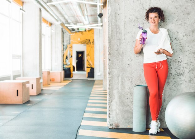 Mujer apta con la botella de agua y el teléfono móvil que se inclinan en la pared en gimnasio