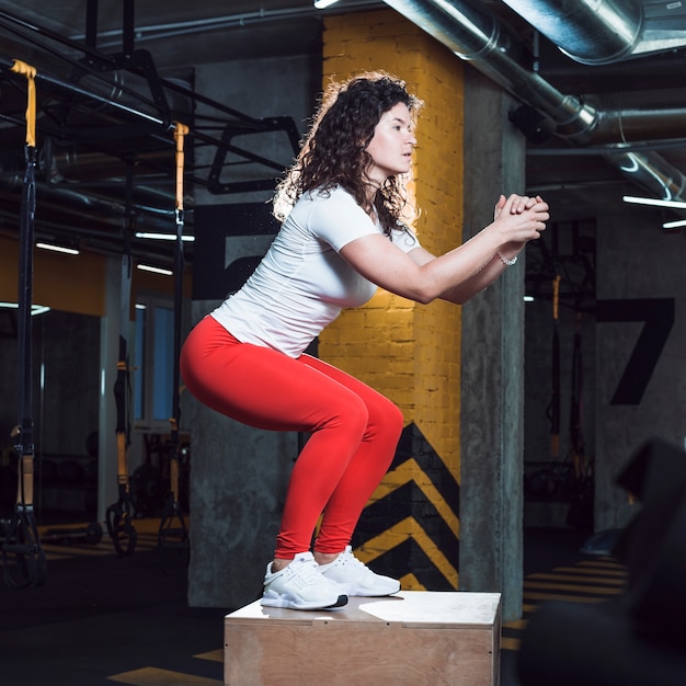 Mujer apta que hace posición en cuclillas en la caja de madera en gimnasio