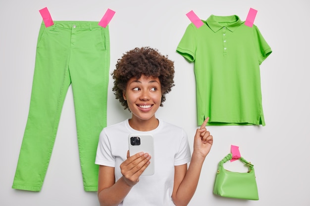 Mujer apunta a casual camiseta verde vende ropa innecesaria limpia armario tiene teléfono móvil hace compras en línea anuncia artículos poses interiores