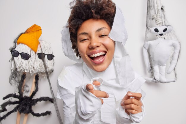 La mujer apunta directamente a la cámara recoge los accesorios para el carnaval disfruta de la época de octubre y las vacaciones espeluznantes viste un disfraz de fantasma sonríe ampliamente posa interior en blanco