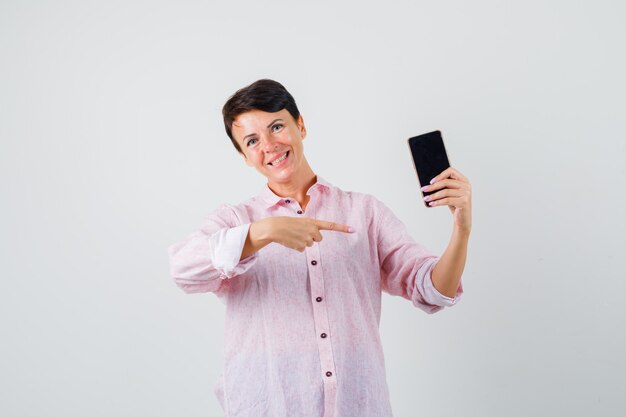 Mujer apuntando al teléfono móvil en camisa rosa y mirando alegre, vista frontal.