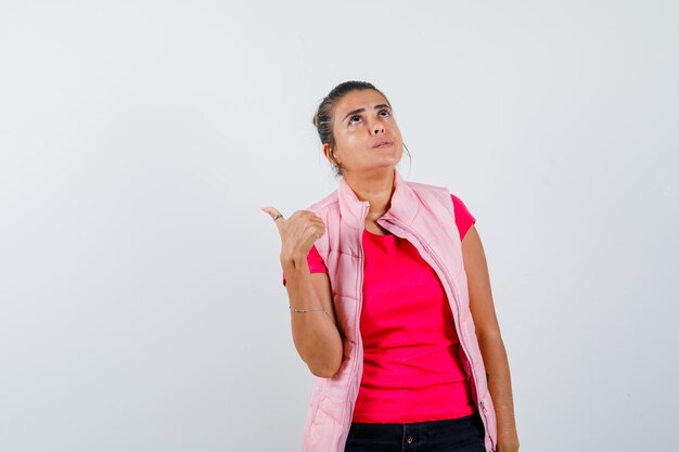 Mujer apuntando hacia atrás con el pulgar en camiseta, chaleco y mirando pensativo