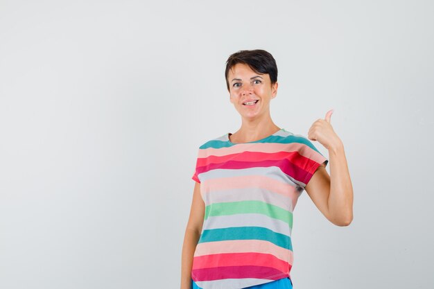 Mujer apuntando hacia atrás con el pulgar en camiseta a rayas y mirando esperanzado, vista frontal.