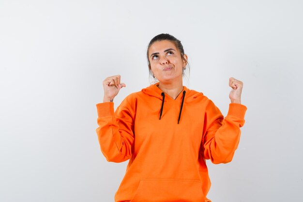 Mujer apuntando hacia atrás con los pulgares en sudadera con capucha naranja y mirando vacilante