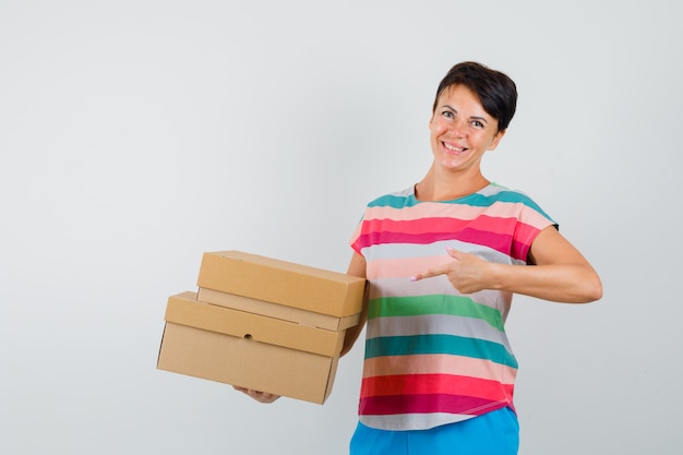 Foto gratuita mujer apuntando a cajas de cartón con camiseta a rayas, pantalones y aspecto alegre. vista frontal.