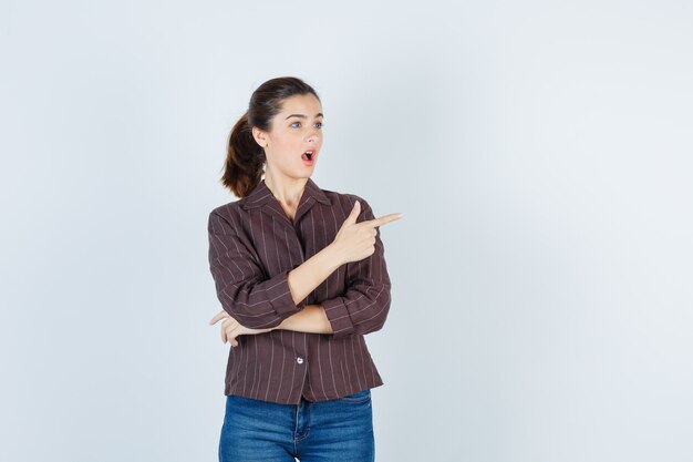 mujer apuntando a la derecha en camisa, jeans y mirando sorprendida. vista frontal.