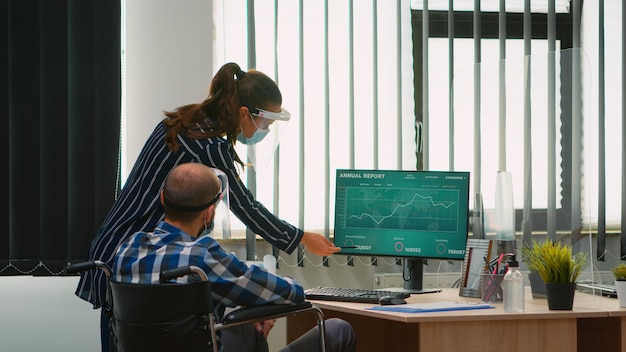 Mujer apuntando en el escritorio hablando con un colega discapacitado en la nueva oficina normal frente a la computadora. Equipo de expertos financieros que trabajan en equipo analizando gráficos económicos respetando la distancia social.