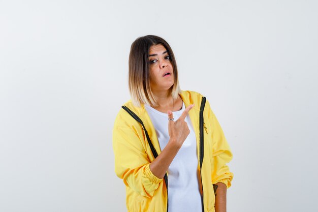 Mujer apuntando a la esquina superior derecha en camiseta, chaqueta y mirando vacilante. vista frontal.