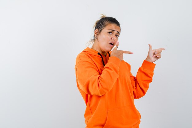 Mujer apuntando hacia el lado derecho con sudadera con capucha naranja y mirando confundida