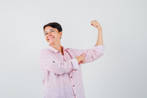 Mujer apuntando a los músculos del brazo con camisa rosa y luciendo orgullosa, vista frontal.