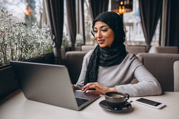 Mujer árabe en hijab dentro de un café trabajando en una computadora portátil