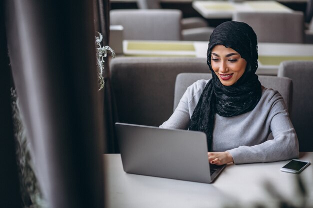 Mujer árabe en hijab dentro de un café trabajando en una computadora portátil