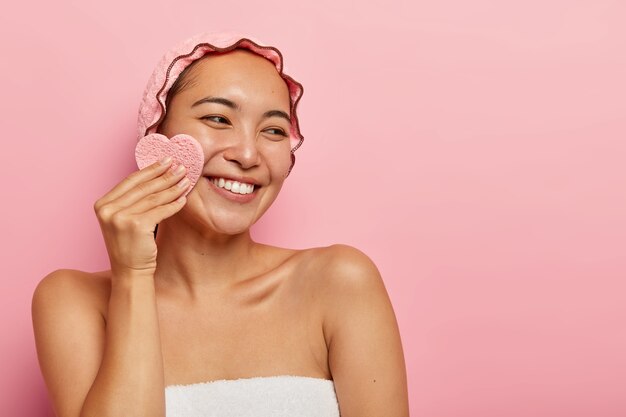 Una mujer asiática alegre y de aspecto agradable se limpia la cara con una esponja cosmética, se quita el maquillaje, se ve del lado derecho, usa un casco protector impermeable, tiene una sonrisa tierna, dientes blancos