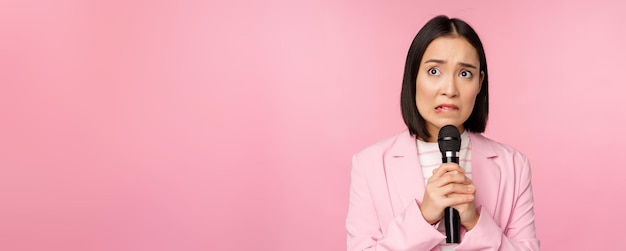 Mujer asiática ansiosa con traje hablando en público dando un discurso con micrófono en una conferencia mirando asustada de pie sobre un fondo rosa
