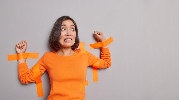 Mujer asiática asustada con cabello oscuro pegado con cintas adhesivas a la pared gris muerde los labios tiene expresión nerviosa aislada sobre la pared gris