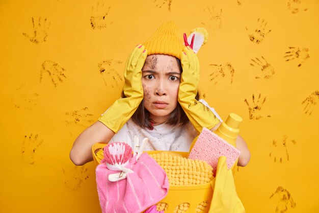 Mujer asiática aterrorizada con la cara sucia ocupada haciendo quehaceres domésticos lleva guantes de goma protectores posa cerca de la canasta de ropa aislada sobre la pared amarilla