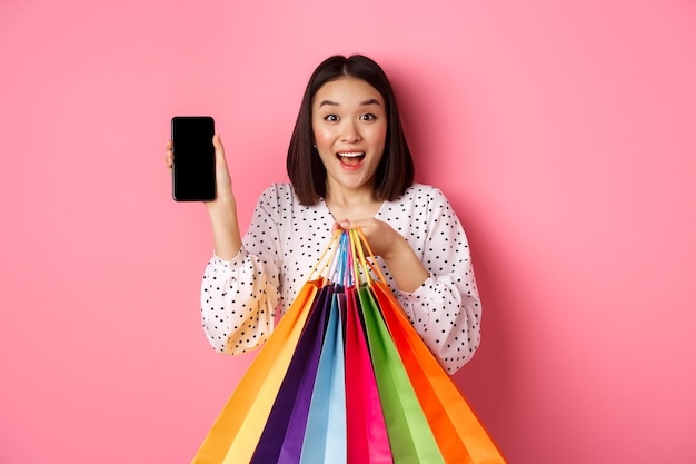 Mujer asiática atractiva que muestra la aplicación del teléfono inteligente y bolsas de compras comprando en línea a través de la aplicación standi ...